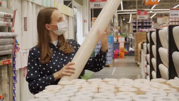 Una mujer con una máscara protectora compra papel pintado en una ferretería. El concepto de reparación y contratación. — Vídeos de Stock