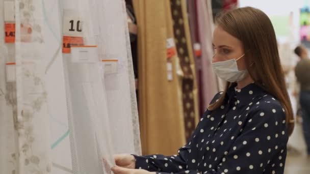 Молода жінка в захисній масці вибирає нові штори в магазині. Зразки штор висять в магазині . — стокове відео