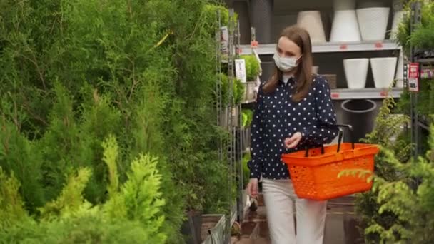 Jonge vrouw met een beschermend masker kiest en koopt een tuya in een tuincentrum in het vroege voorjaar. — Stockvideo