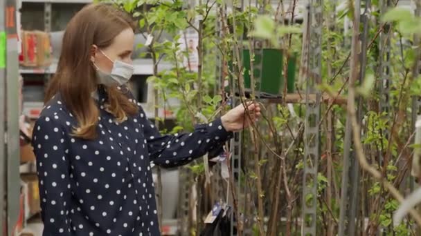 Una mujer con una máscara protectora escoge un árbol frutal en un centro de jardinería. — Vídeos de Stock