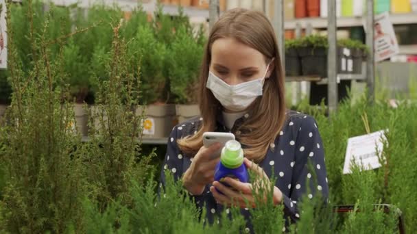 医療用マスクの女性は、スマートフォンを使用して花や植物の農薬を選択します — ストック動画