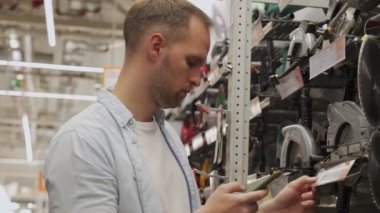 Adam bir alet dükkanında akıllı telefon kullanarak dairesel bir testere seçiyor. Erkek bir hırdavatçıdan ürün seçer..