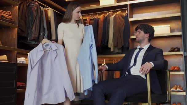 La vendedora sonríe y ofrece las camisas a un joven hombre de negocios guapo moderno en una tienda de disfraces — Vídeos de Stock