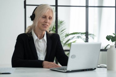 Elderly lady in a headset talks on a video conference, conducts a video conference using a laptop in the office