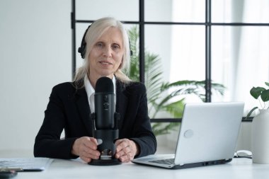 Elderly woman records a podcast on her laptop with headphones and a microphone