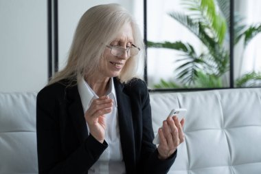 Elderly woman sitting on the couch, smiling while using her phone, celebrating success