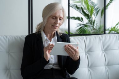 Elderly woman uses a digital tablet while sitting on the couch at home. The use of technology by older people.
