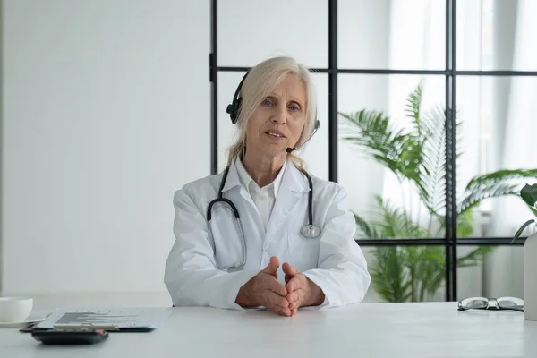 Elderly female doctor in a white medical coat, looking at the camera and talking with headphones, sitting in a hospital office. — Foto de Stock
