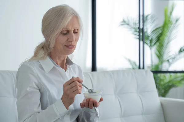 Elderly white-haired woman eats yogurt sitting on the couch — 图库照片