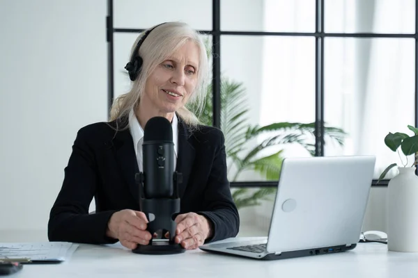 Elderly woman records a podcast on her laptop with headphones and a microphone — Foto de Stock