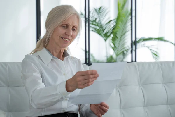 Older woman reading a letter sitting on a sofa in the living room. Overjoyed mature woman with glasses reads the good news in a letter — 图库照片