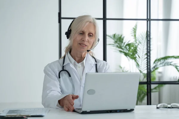 Elderly female doctor with a stethoscope, wearing a lab coat and headset, communicates by video call with a patient via a laptop — Foto de Stock