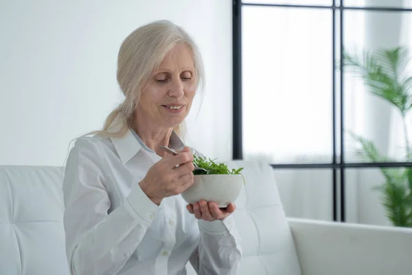 Mature white-haired woman sits on the couch and eats a vegetable salad — 图库照片