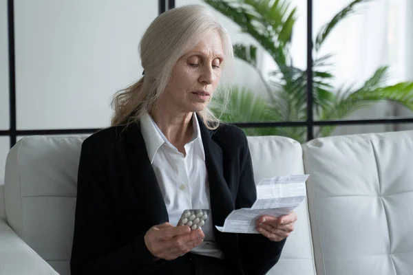 Gray haired elderly woman reads the instructions before taking her medication at home. Grandmother sitting on the couch reads the instructions for the use of tablets — Foto de Stock