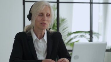 Elderly lady in a headset talks on a video conference, conducts a video conference using a laptop in the office