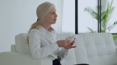 Elderly woman is sitting on the sofa, drinking coffee or tea and looking out the window