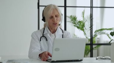 Elderly female doctor with a stethoscope, wearing a lab coat and headset, communicates by video call with a patient via a laptop