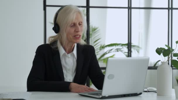 Elderly lady in a headset talks on a video conference, conducts a video conference using a laptop in the office — Vídeo de stock