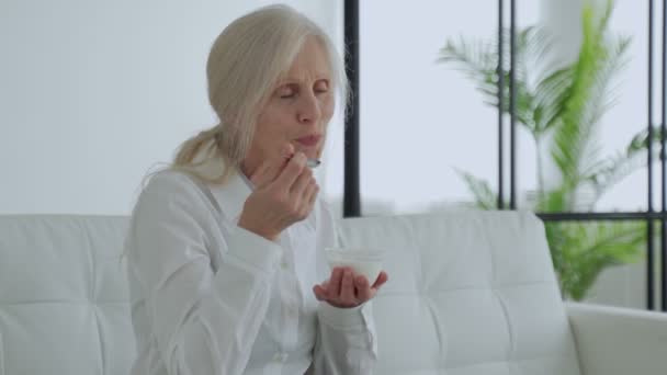 Elderly white-haired woman eats yogurt sitting on the couch — Vídeo de stock