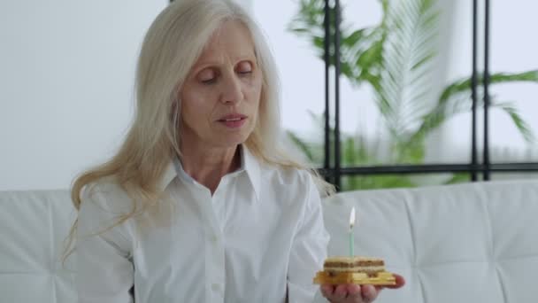 Lonely birthday. A sad depressed elderly white-haired woman celebrates alone by blowing on a cake. — Vídeo de stock