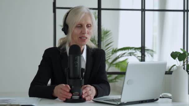 Elderly woman records a podcast on her laptop with headphones and a microphone — Video