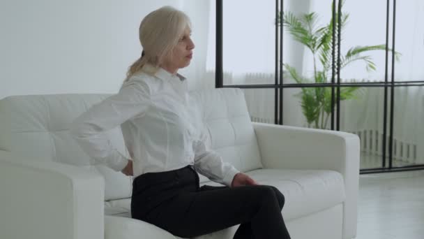 An elderly woman suffering from back pain. A middle-aged woman gets up from the sofa and experiences back pain — Vídeo de stock