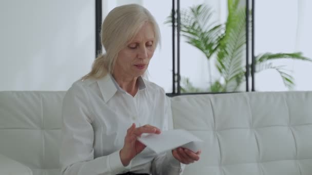 Older woman reading a letter sitting on a sofa in the living room. Overjoyed mature woman with glasses reads the good news in a letter — Wideo stockowe