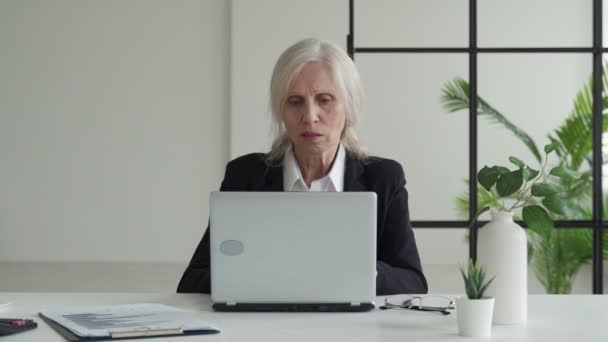 Elderly business woman works in an office with a laptop and looks at the camera — Stockvideo