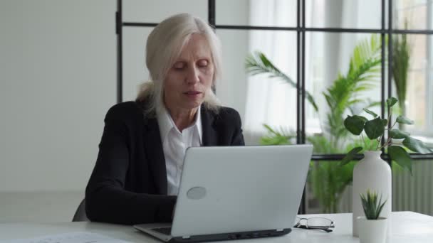 Elderly gray-haired woman uses a laptop comes up with an idea. Businesswoman working in a modern office. — Αρχείο Βίντεο