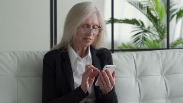 Elderly woman sitting on the couch, smiling while using her phone, celebrating success — Stock video