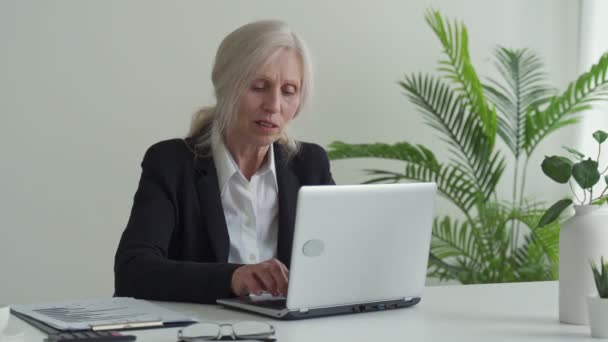 Happy mature woman celebrates online victory with a laptop while sitting at her workplace in the office — Video