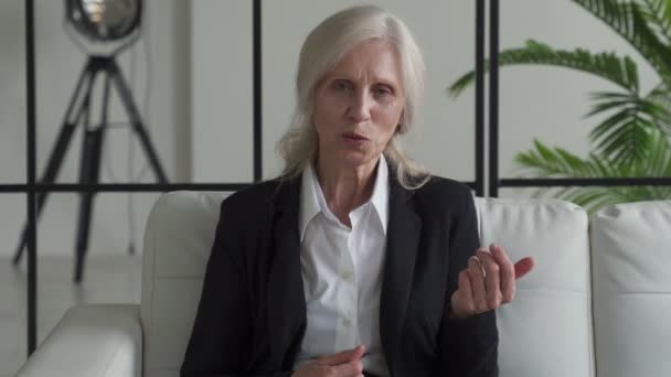 An elderly female businesswoman sitting on the couch. Mature woman looks at the camera, gestures and talks online, during a video call — Vídeo de Stock