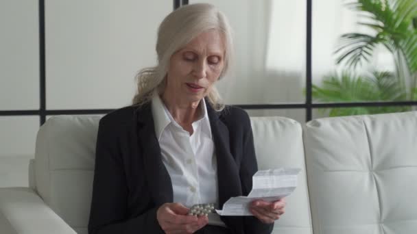 Літня жінка з сивим волоссям читає інструкцію, перш ніж приймати ліки вдома. Бабуся, що сидить на дивані, читає інструкцію про використання табличок. — стокове відео