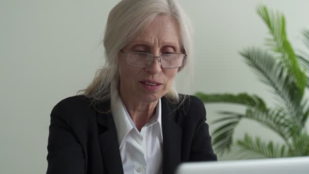 Втомилася розчарована зріла сіроволоса жінка, знімає окуляри, відпочиває від роботи на ноутбуці, масажує голову рукою — стокове відео