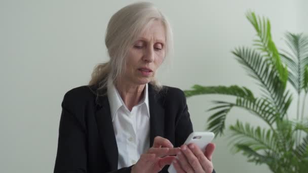 Smiling elderly woman holding a phone, using mobile apps, looking at the screen, writing a message on a mobile phone while sitting on the couch — Vídeo de stock