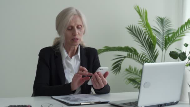 Excited elderly business woman enjoys the good news on her mobile phone while sitting at her desk in the office — Stockvideo