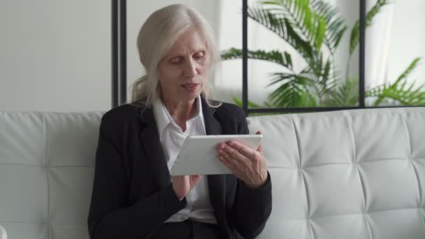 Elderly woman uses a digital tablet while sitting on the couch at home. The use of technology by older people. — Stok video