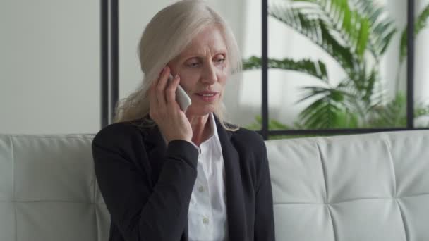 Portrait of an elderly woman in a business suit, sitting on a sofa and talking on the phone with a smile. An elderly woman is talking on a mobile phone. The concept of communication and retirement — Stockvideo