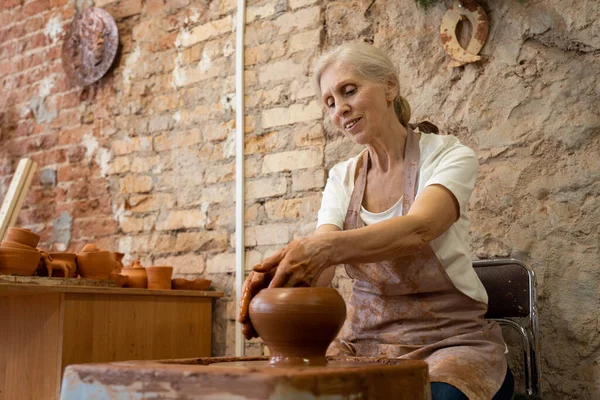 Anciana alfarera trabajando en rueda de cerámica mientras está sentada en su taller — Foto de Stock