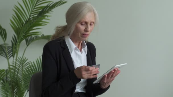 Mujer mayor en un traje de negocios se sienta en una silla y utiliza una tableta — Vídeo de stock