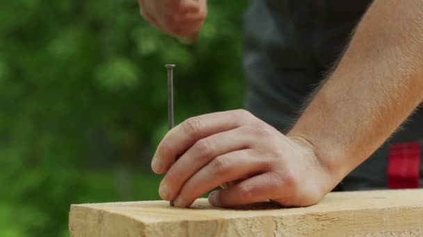 Плотник вбивает гвоздь в деревянную доску, стоя снаружи — стоковое видео