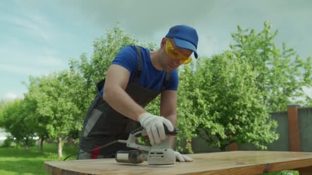 Carpintero masculino con gafas de seguridad y guantes pule una mesa de madera con una rectificadora en el taller — Vídeos de Stock