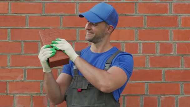 A man wearing a cap and gloves shows a brick to the camera in a construction concept — Stock Video