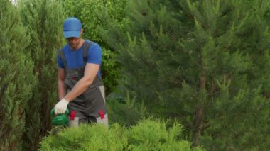 Koruyucu kıyafetli erkek bahçıvan elektrikli budayıcı ile çalıları buduyor.