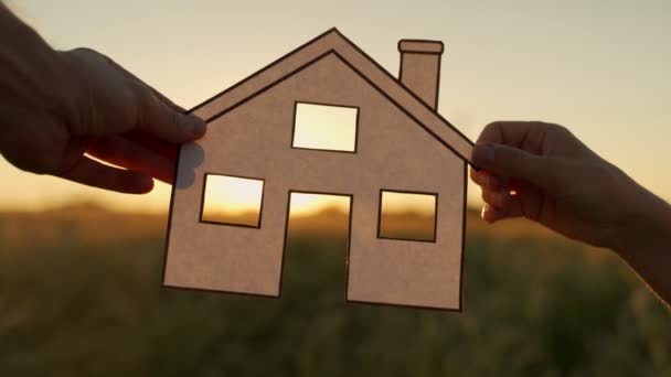 A család papírházat tart napnyugtakor, a nap kisüt az ablakon. Kézi tartó papír vágott ház szimbólumok naplementekor — Stock videók