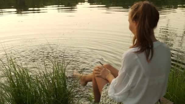 Junge Frau auf der Anklagebank, die mit nackten Beinen im See planscht. Frauenfüße im Wasser lassen Frische spüren. — Stockvideo