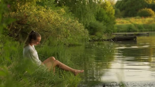 Jovem mulher sentada na doca salpicando pernas nuas no lago. Pés de mulher na água mergulha sentindo frescura. — Vídeo de Stock