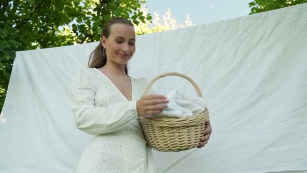 Young woman stands in front of an apple tree and holds a basket with laundry in her hands and is drying bed linen in the fresh wind on a rope. — Stock Video