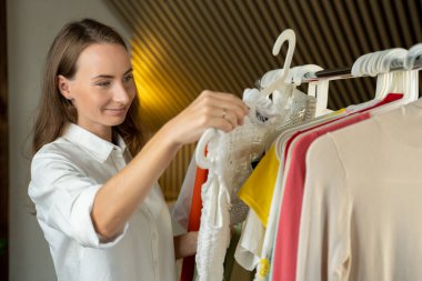 Gömlekli genç bir kadın askı önünde duruyor ve iş için ya da yürüyüş için bir kıyafet seçmeye çalışıyor..