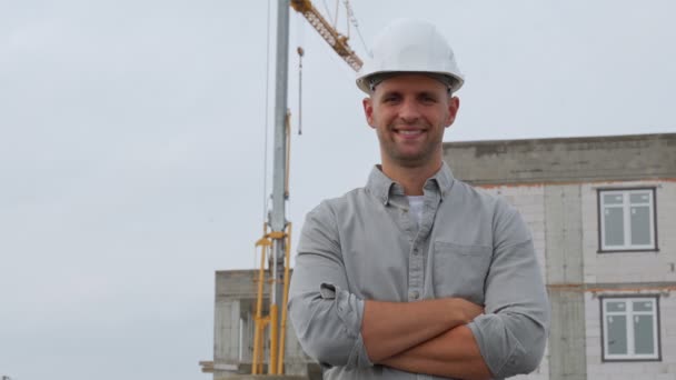 Architecte dans un casque protecteur des constructeurs sur fond de maison en construction, souriant avec les bras croisés, regarde la caméra. — Video
